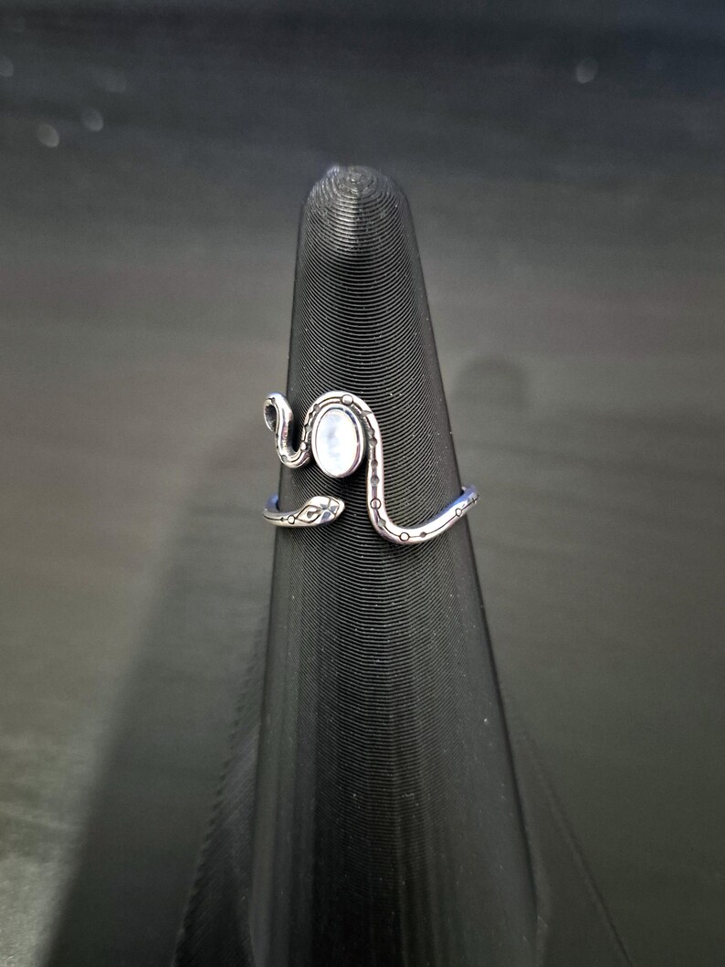 May include: A silver-toned metal ring shaped like a coiled snake with a smooth, oval-shaped white stone set in the center. The band features etched detailing along the body of the snake, including small circular markings and a defined head shape.