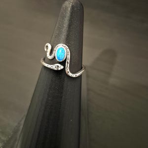 May include: A silver-toned metal ring shaped like a coiled snake with a smooth, oval-shaped bright blue stone set in the center. The snake body features etched black line details and small circular patterns along the band, wrapping around the finger.