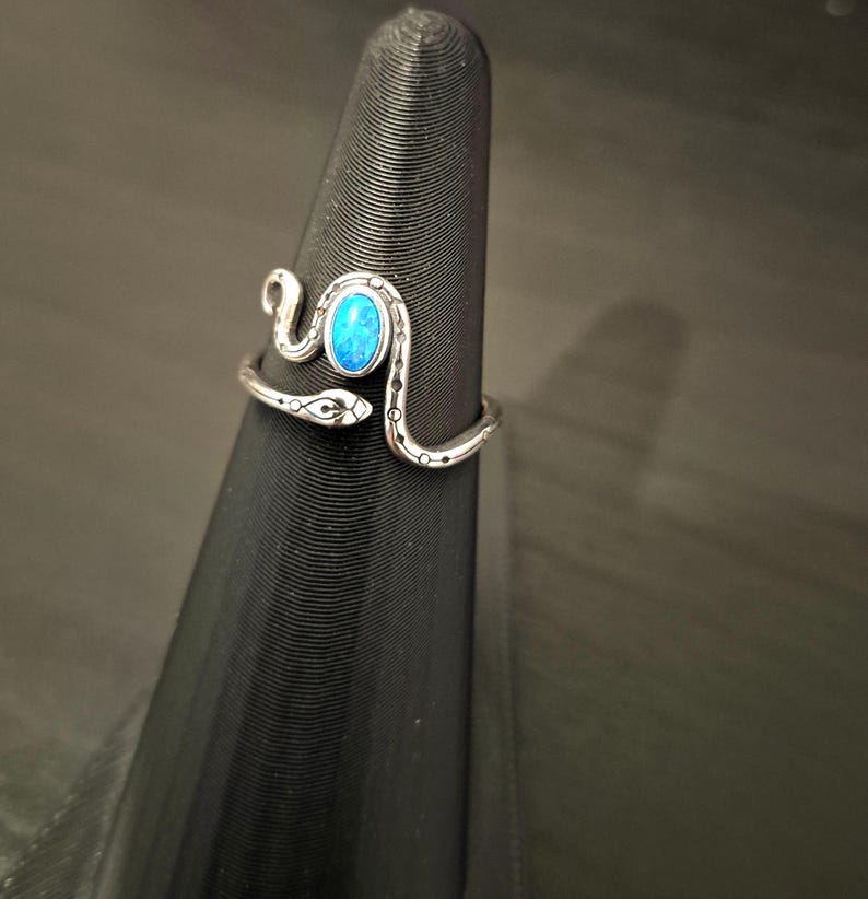 May include: A silver-toned metal ring shaped like a coiled snake with a smooth, oval-shaped bright blue stone set in the center. The snake body features etched black line details and small circular patterns along the band, wrapping around the finger.