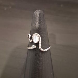 May include: A silver-toned metal ring shaped like a coiled snake with an open design. The snake features engraved scale patterns along its body and a smooth, oval-shaped white iridescent stone set in the center of the head area.