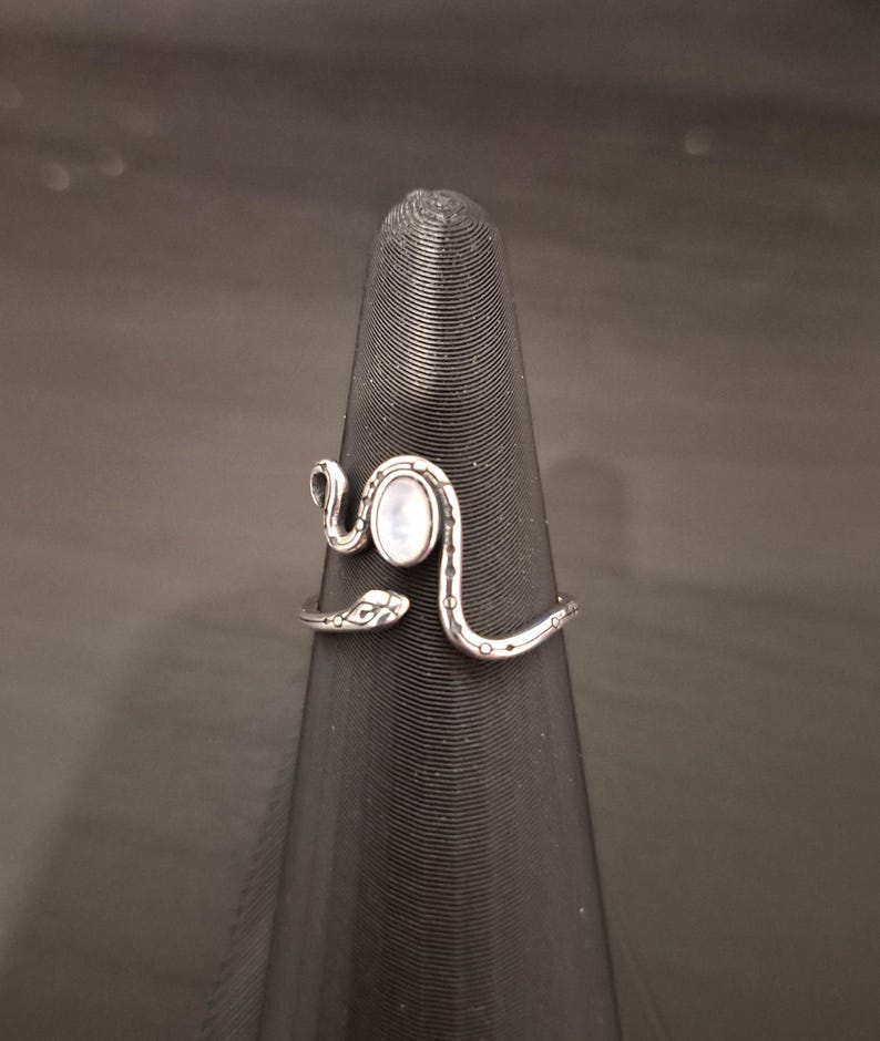 May include: A silver-toned metal ring shaped like a coiled snake with an open design. The snake features engraved scale patterns along its body and a smooth, oval-shaped white iridescent stone set in the center of the head area.