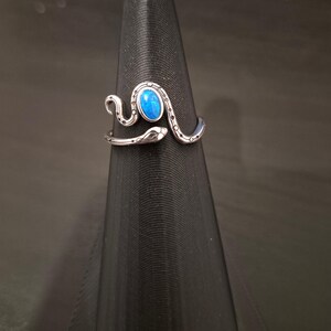 May include: A silver-toned metal ring shaped like a coiled snake with a textured, dotted pattern along its body. The snake features an oval-shaped, vibrant blue cabochon stone set prominently on its back, with the head and tail ends overlapping.