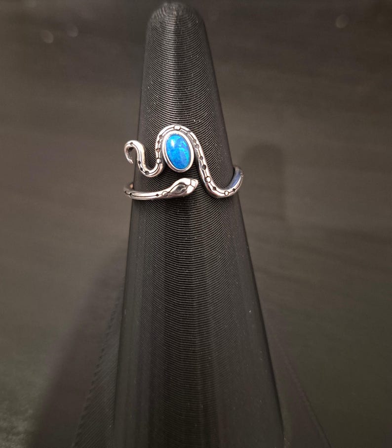 May include: A silver-toned metal ring shaped like a coiled snake with a textured, dotted pattern along its body. The snake features an oval-shaped, vibrant blue cabochon stone set prominently on its back, with the head and tail ends overlapping.