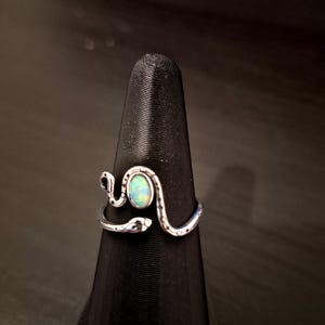 May include: A silver-toned ring shaped like a coiled snake with a textured body and a smooth, oval-shaped iridescent opal stone set in the center. The snake's head and tail wrap around the finger, meeting at the front to frame the central gemstone.
