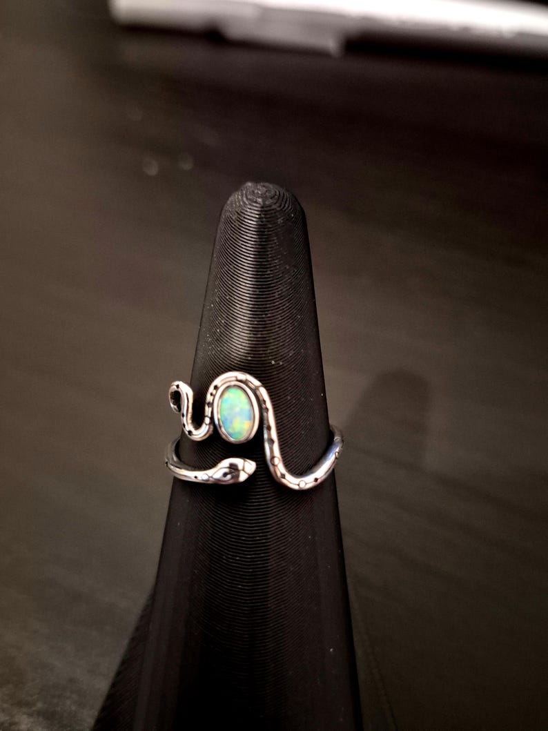 May include: A silver-toned ring shaped like a coiled snake with a textured body and a smooth, oval-shaped iridescent opal stone set in the center. The snake's head and tail wrap around the finger, meeting at the front to frame the central gemstone.
