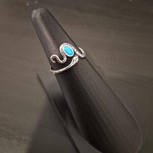 May include: A silver-toned metal ring shaped like a coiled snake with an oval-shaped, iridescent blue stone set into the head. The snake body features etched detailing along the band, creating a textured appearance that wraps around the finger.