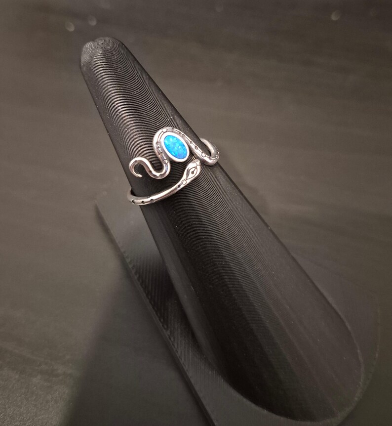 May include: A silver-toned metal ring shaped like a coiled snake with an oval-shaped, iridescent blue stone set into the head. The snake body features etched detailing along the band, creating a textured appearance that wraps around the finger.