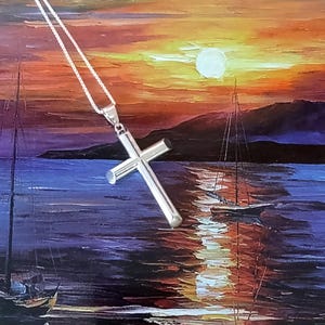 May include: A silver cross pendant on a silver chain. The cross is a simple, polished design. The background is a painting of a sunset over the ocean with sailboats.
