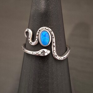May include: A silver-toned metal ring shaped like a coiled snake with a textured, dotted pattern along its body. The center of the ring features an oval-shaped, vibrant blue stone with a shimmering, iridescent finish set within the snake's curve.