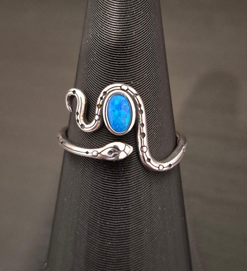 May include: A silver-toned metal ring shaped like a coiled snake with a textured, dotted pattern along its body. The center of the ring features an oval-shaped, vibrant blue stone with a shimmering, iridescent finish set within the snake's curve.