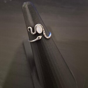 May include: A silver-toned open ring shaped like a coiled snake with a smooth, oval-shaped white stone set in the center. The metal band features engraved dot and line details along the body of the snake, which wraps around the finger area.