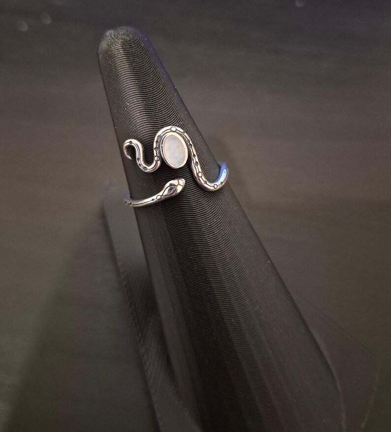 May include: A silver-toned open ring shaped like a coiled snake with a smooth, oval-shaped white stone set in the center. The metal band features engraved dot and line details along the body of the snake, which wraps around the finger area.