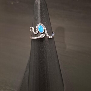 May include: A silver-toned metal ring shaped like a coiled snake with a textured, patterned body. The snake's head is positioned at one end of the open band, and a small, oval-shaped, vibrant blue stone is set prominently in the center of the coil.