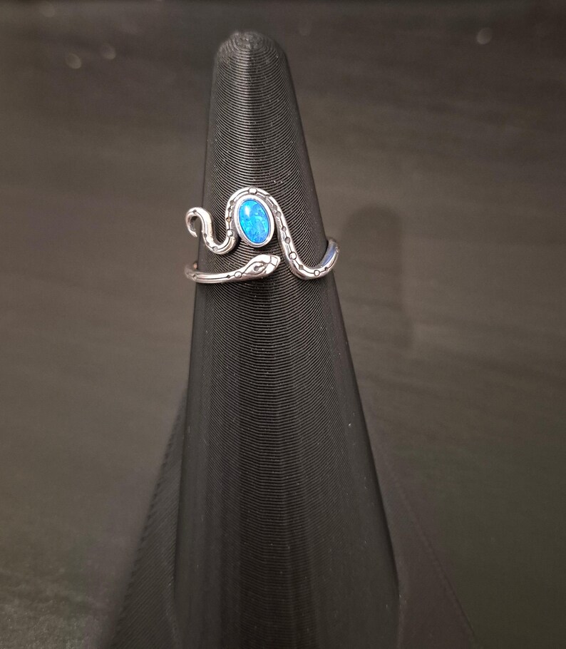 May include: A silver-toned metal ring shaped like a coiled snake with a textured, patterned body. The snake's head is positioned at one end of the open band, and a small, oval-shaped, vibrant blue stone is set prominently in the center of the coil.