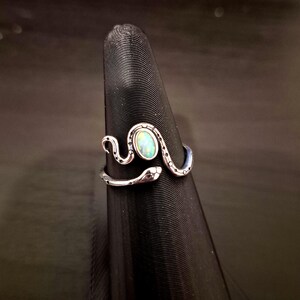 May include: A silver-toned metal ring shaped like a coiled snake with a textured, dotted pattern along its body. The snake's head and tail meet at the front, framing a central, oval-shaped iridescent stone that displays flashes of blue and green.
