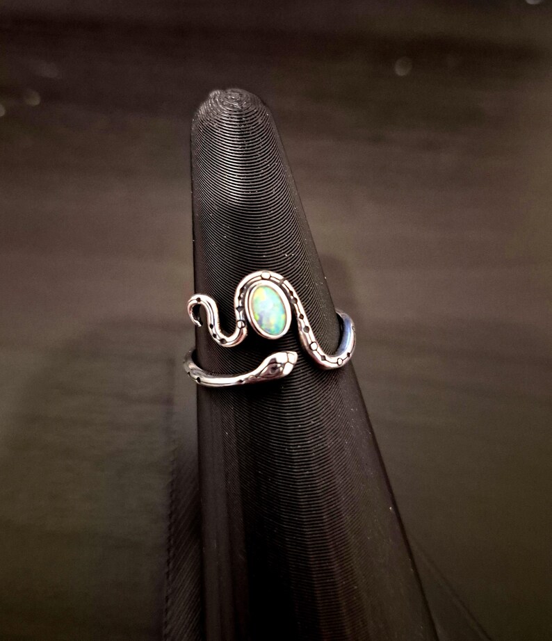May include: A silver-toned metal ring shaped like a coiled snake with a textured, dotted pattern along its body. The snake's head and tail meet at the front, framing a central, oval-shaped iridescent stone that displays flashes of blue and green.