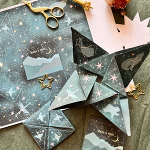 May include: A set of two origami star-shaped paper cards with a dark blue background and gold stars. The cards have the text "Make a wish" printed on them. The cards are surrounded by other craft supplies, including a pair of gold scissors, a gold pen, and a gold star-shaped paper clip.