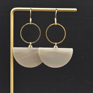 May include: A pair of dangle earrings with a gold-colored circular top and a semi-circular, pearlescent, gray-white bottom. The earrings are displayed on a gold-colored stand against a black background.