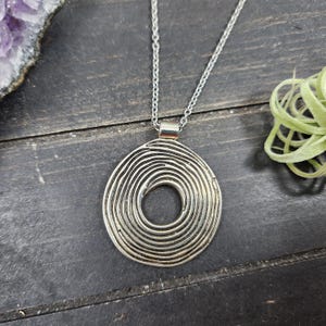 May include: Silver pendant necklace with a spiral design. The pendant is a circular shape with concentric lines. The necklace hangs from a fine chain. The image shows the necklace on a dark wooden surface, with a hint of amethyst and a small plant.