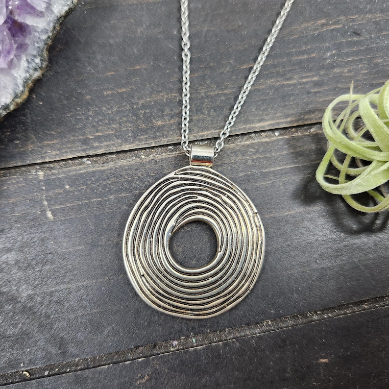 May include: Silver pendant necklace with a spiral design. The pendant is a circular shape with concentric lines. The necklace hangs from a fine chain. The image shows the necklace on a dark wooden surface, with a hint of amethyst and a small plant.