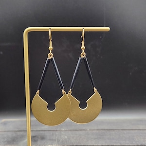 May include: A pair of gold earrings with black geometric shapes. The earrings are in the shape of a crescent moon with a small circle in the center.