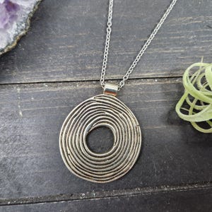 May include: Silver pendant necklace with a spiral design. The circular pendant has concentric lines forming a swirl. The necklace has a fine chain and a simple clasp. The background is a dark wood surface.