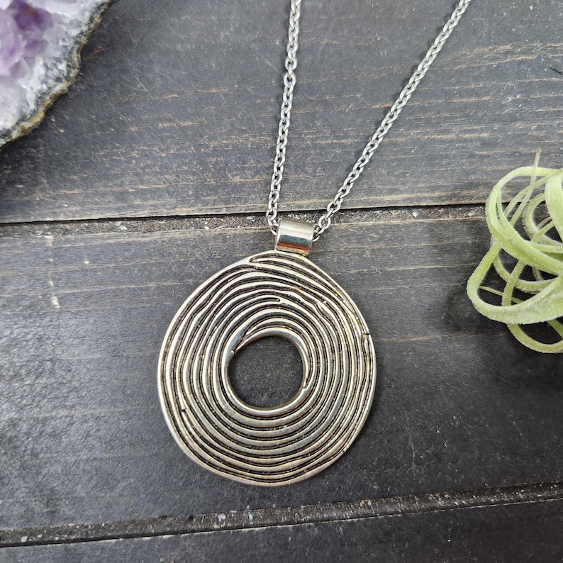 May include: Silver pendant necklace with a spiral design. The circular pendant has concentric lines forming a swirl. The necklace has a fine chain and a simple clasp. The background is a dark wood surface.