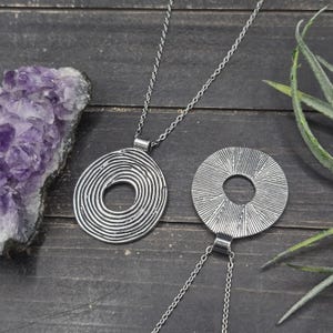 May include: Two silver pendant necklaces. One pendant has a spiral design, the other a radial pattern. They are displayed on a dark wooden surface, with a purple crystal and greenery in the background.