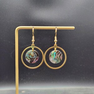 May include: A pair of dangle earrings featuring a circular gold-tone frame with a central iridescent abalone shell disc. The earrings hang from gold-tone fishhook wires, showcasing a natural swirling pattern of green, blue, and purple hues.
