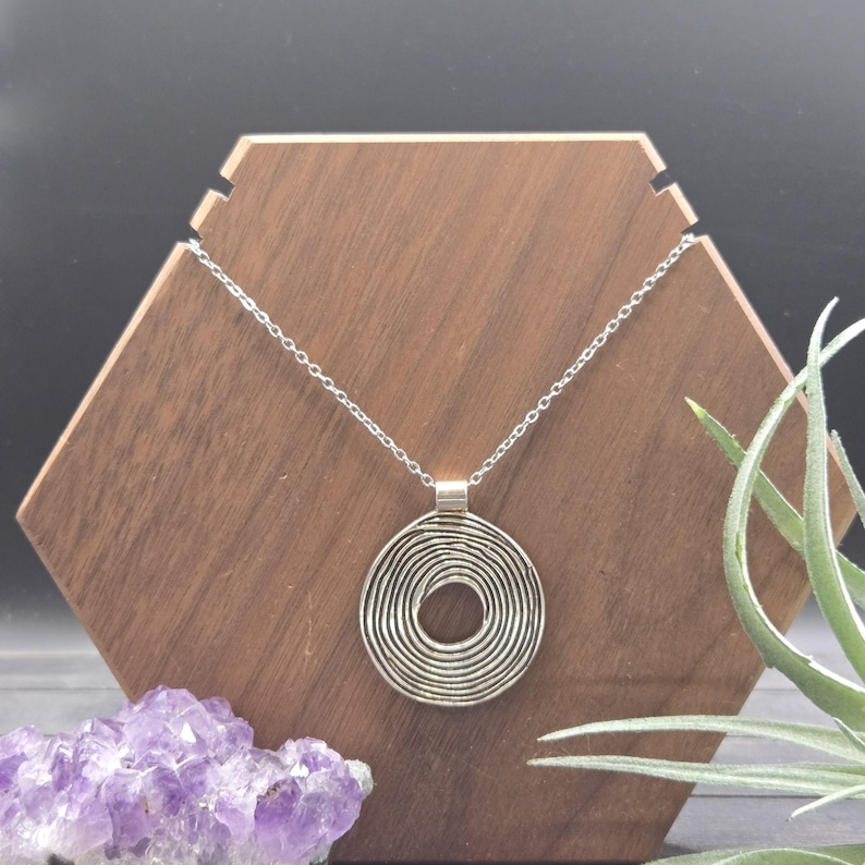 May include: A silver pendant necklace with a spiral design. The pendant hangs from a delicate chain. It is displayed on a wooden hexagonal stand. A cluster of purple amethyst crystals and greenery are in the foreground.