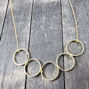May include: A gold necklace with five interlocking circles. The necklace is on a dark wood background.