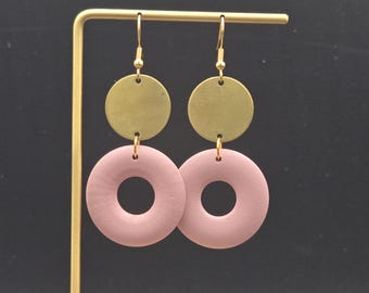 May include: A pair of dangle earrings featuring a brass disc above a pink donut-shaped element. The earrings hang from gold-colored ear wires. The background is a dark gray.