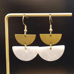 May include: A pair of gold-tone and white half-circle dangle earrings. The top half-circle is a solid gold colour, while the bottom half-circle is white with a marbled effect. The earrings hang from gold-coloured fish hook ear wires.