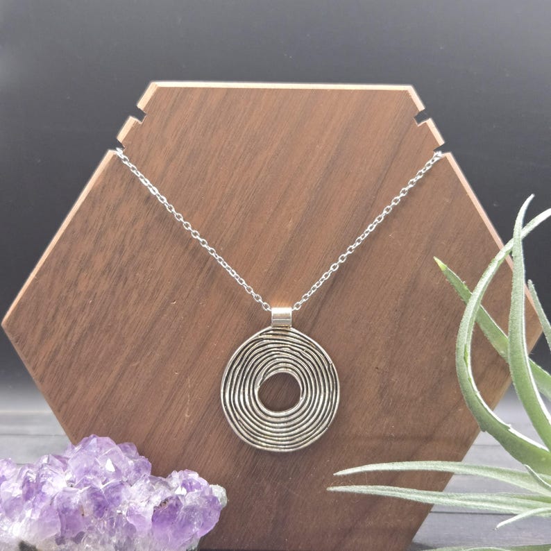 May include: A silver pendant necklace featuring a spiral design. The pendant is suspended from a fine chain. The necklace is presented on a wooden hexagonal stand, with a purple amethyst crystal and a green plant in the background.