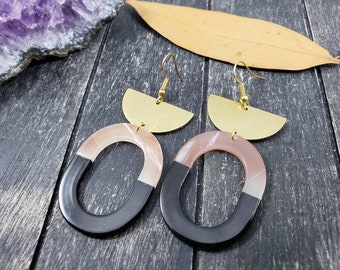May include: A pair of statement earrings featuring a black and pink oval shape with a gold half-circle accent. The earrings have a modern geometric design.