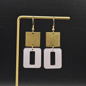 May include: A pair of gold-tone square earrings with a textured finish, connected to white, open-rectangle pendants. The earrings are displayed on a gold-colored stand against a black background. The jewelry is designed for everyday wear.