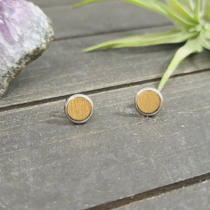 May include: A pair of small, round, wooden earrings with a silver metal setting. The earrings are a light brown color and have a wood grain pattern.
