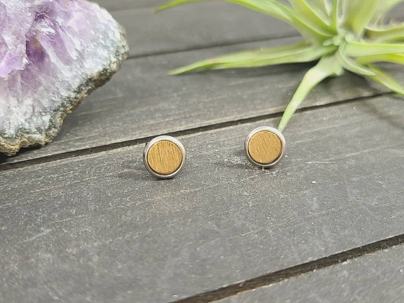 May include: A pair of small, round, wooden earrings with a silver metal setting. The earrings are a light brown color and have a wood grain pattern.