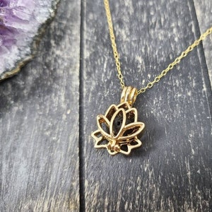 May include: Gold lotus flower pendant necklace with a small black lava stone inside. The necklace is on a wooden surface.