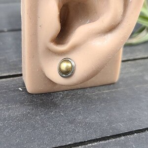Puede incluir: Primer plano de un pendiente de bola dorada en una oreja de maniqu&iacute;. El pendiente es de metal y tiene un acabado liso y brillante.