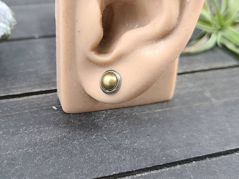Puede incluir: Primer plano de un pendiente de bola dorada en una oreja de maniqu&iacute;. El pendiente es de metal y tiene un acabado liso y brillante.
