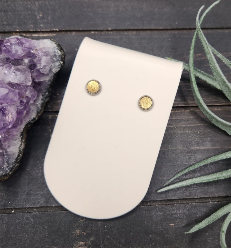 May include: A pair of gold and silver stud earrings on a white display card. The earrings are round and have a textured surface.