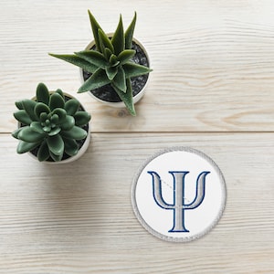 Könnte beinhalten: Ein weißer runder Patch mit einem grauen gestickten griechischen Buchstaben Psi auf einer weißen Holzoberfläche mit zwei grünen Sukkulenten in weißen Töpfen.