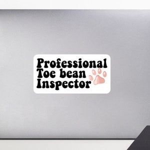 Puede incluir: Un portátil blanco con una pegatina negra que dice "Professional Toe bean Inspector" con una huella de pata rosa.