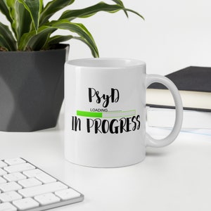 Könnte beinhalten: Weiße Keramiktasse mit einer schwarzen Grafik, auf der "PsyD Loading... in progress" mit einem grünen Fortschrittsbalken steht.