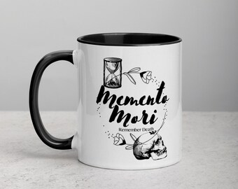 May include: White ceramic coffee mug with a black rim and handle. The mug features a black and white illustration of an hourglass, a skull, flowers, and the text "memento mori" and "Remember Death".