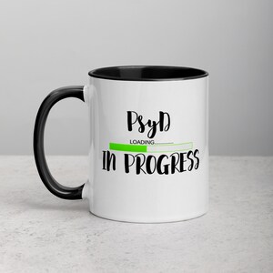 Könnte beinhalten: Weiße Keramik-Kaffeetasse mit schwarzem Rand und Henkel. Die Tasse hat eine schwarze Grafik mit der Aufschrift "PsyD Loading... in progress" und einem grünen Fortschrittsbalken.