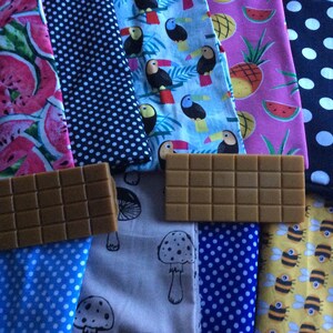 May include: An assortment of colorful fabric swatches with various patterns, including watermelons, toucans, pineapples, polka dots, mushrooms, and bees. Two rectangular, brown, grid-patterned objects are in the foreground.
