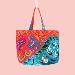 May include: A colorful tote bag with a vibrant paisley pattern. The bag features a combination of pink, orange, blue, and green hues. The handles are red and blue, and the bag is suspended against a pink background.