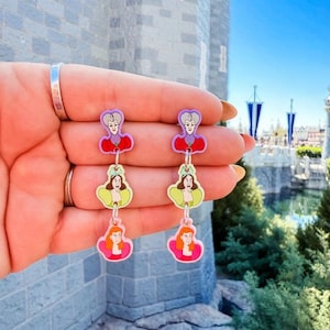 May include: A pair of dangle earrings featuring three different characters from the Disney movie Cinderella. The top charm is the evil stepmother, the middle charm is the fairy godmother, and the bottom charm is Cinderella. The earrings are made of plastic and have a colorful design.